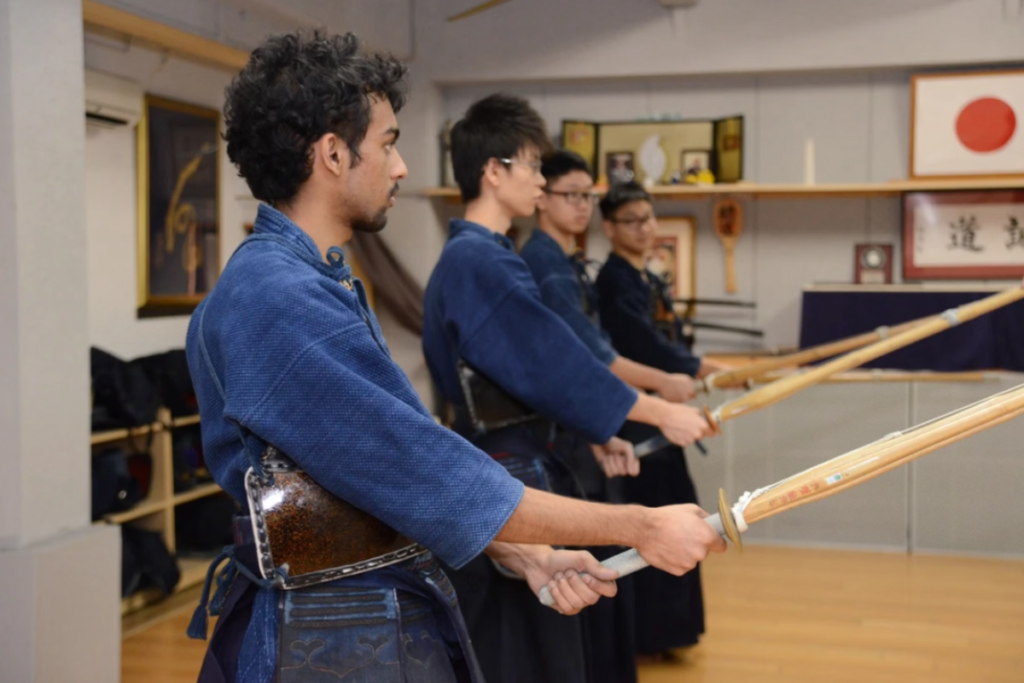 A line of young men in dark blue martial arts practice uniforms standing side by side, holding wooden and bamboo swords during a Kendo class in a Tokyo dojo.