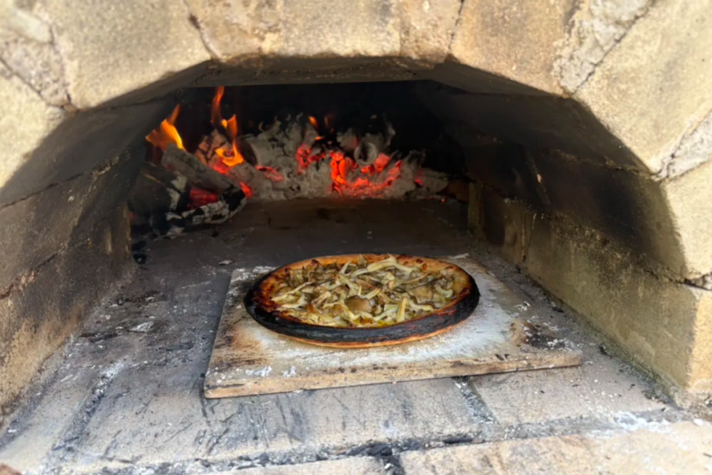 A fresh pizza topped with mushrooms and ingredients bakes inside a traditional, glowing stone oven with burning embers in the Kyoto countryside.