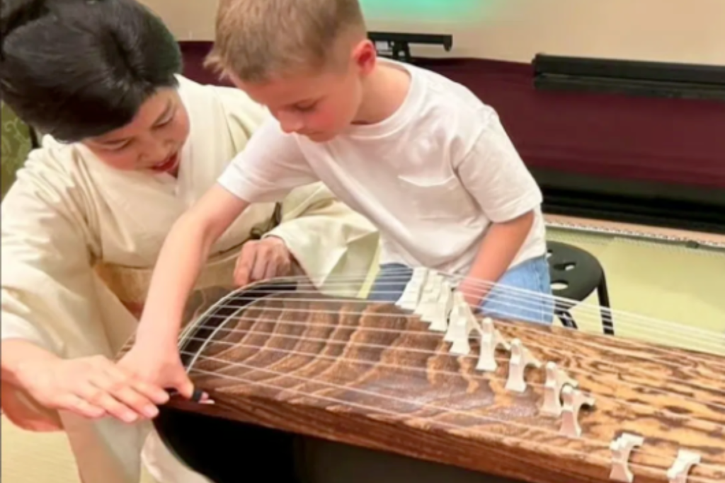 A professional Japanese musician in a kimono guides a young student on how to pluck the strings of a traditional wooden Koto instrument during a private lesson.