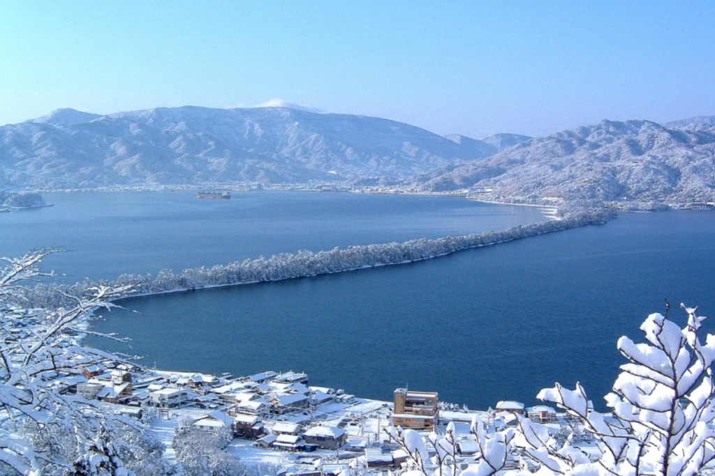 High-angle panoramic view of the snow-covered Amanohashidate sandbar stretching across the blue water of the bay in winter.