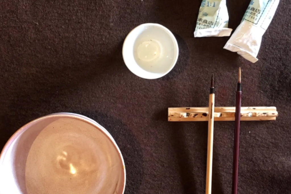 A top-down view of a traditional ceramic Kintsugi bowl alongside two wooden application brushes on a dark studio table.