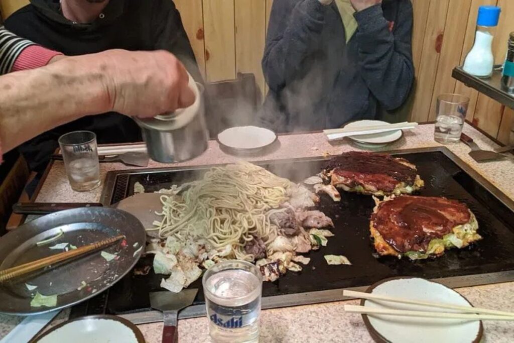 A chef preparing a hot savory cabbage pancake on a steaming iron griddle.