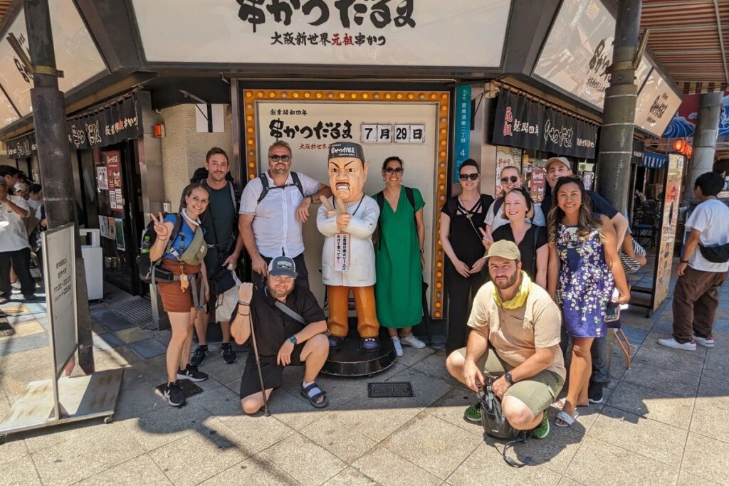 A large group of travelers posing outside a popular restaurant during an Osaka history food tour.
