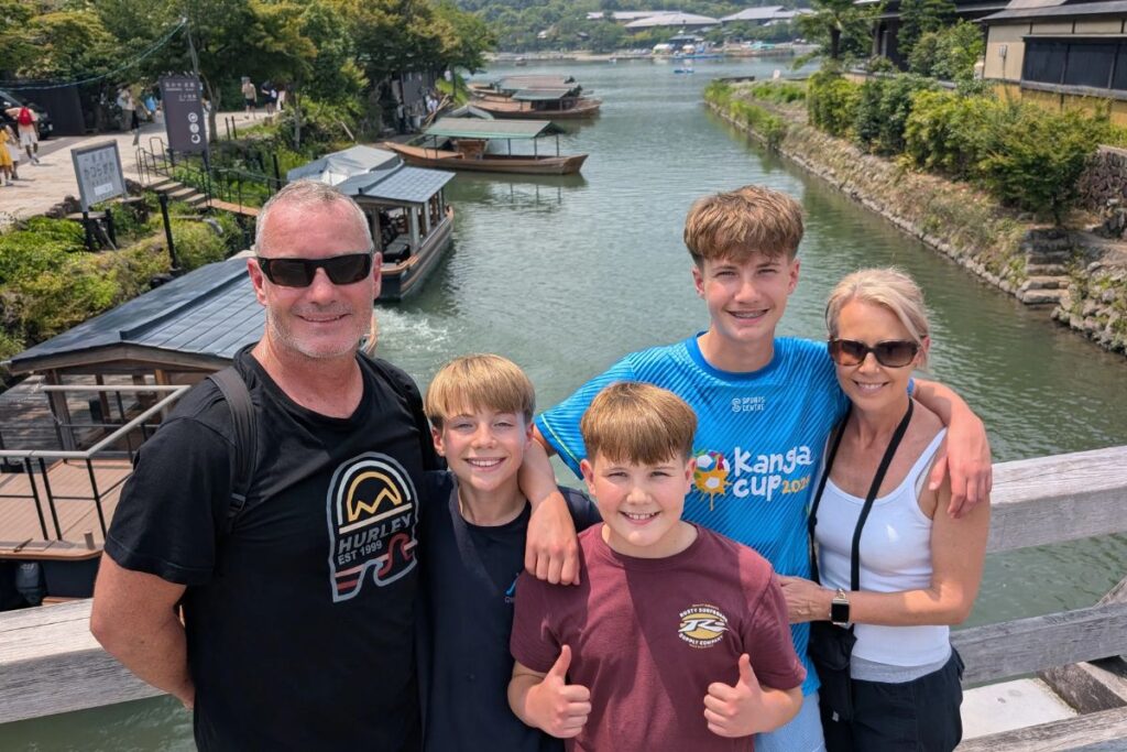 A family of four smiling on a wooden bridge overlooking a scenic river with small boats.