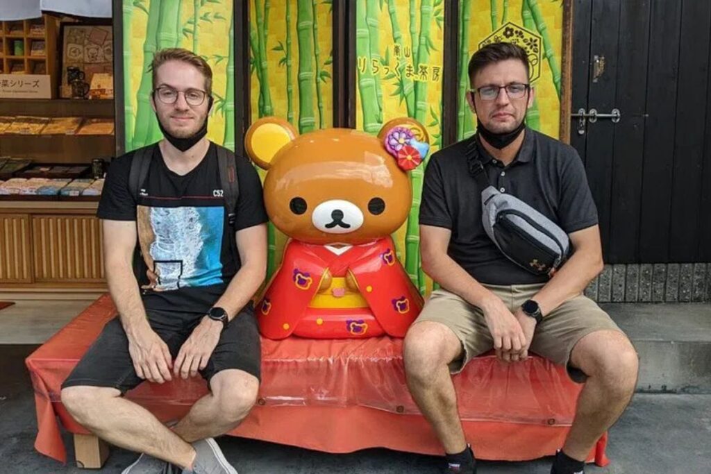 Two men sitting on a red bench next to a cute mascot character statue wearing a traditional outfit.