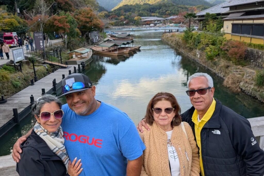 A family of four smiling on a wooden bridge overlooking a scenic river with small boats.