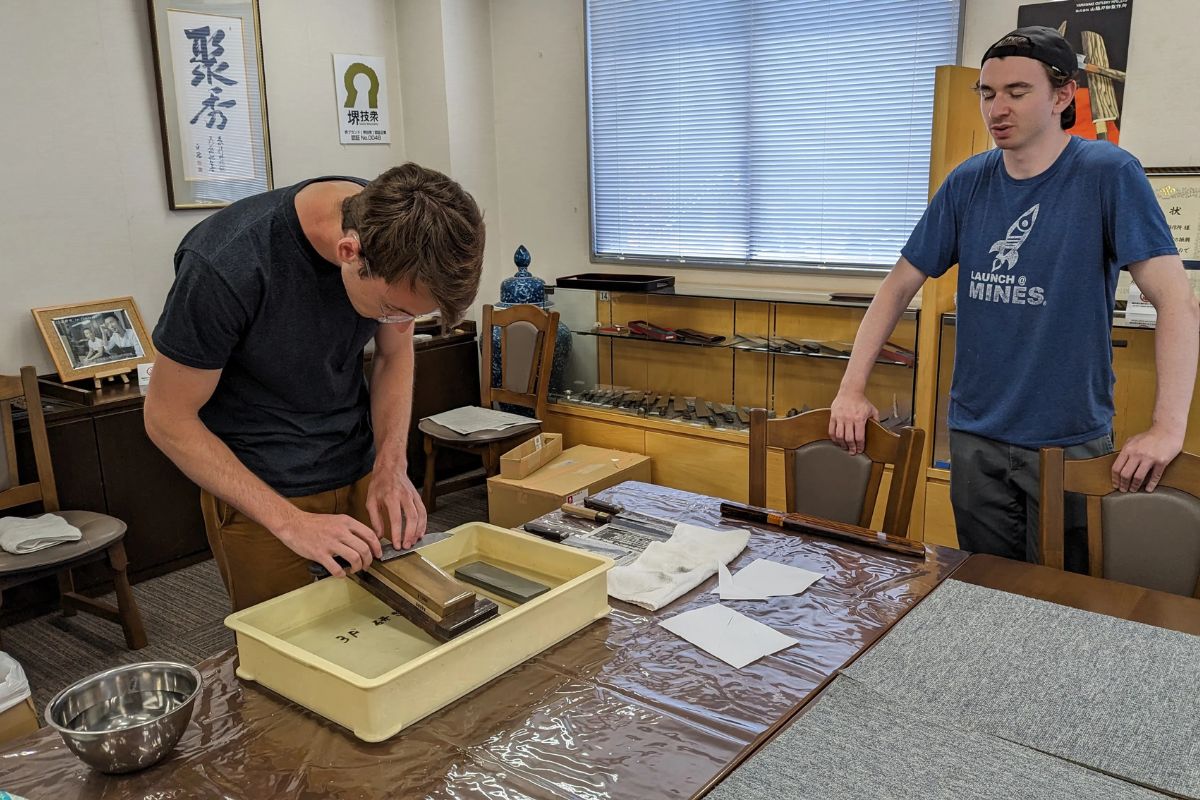 A young man practicing traditional blade sharpening on a wet stone while a friend watches.
