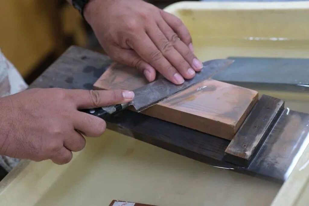 Close-up of an artisan carefully sharpening a steel cooking blade on a wet sharpening stone.