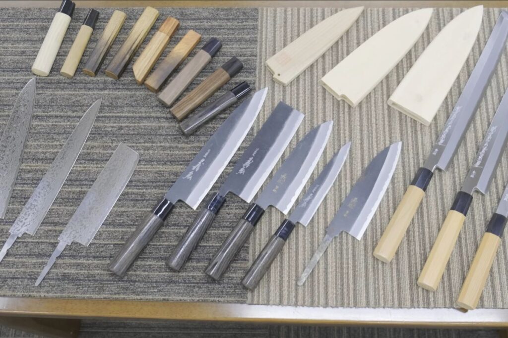 A display table showing various cooking blades, wooden handles, and protective wooden sheaths.