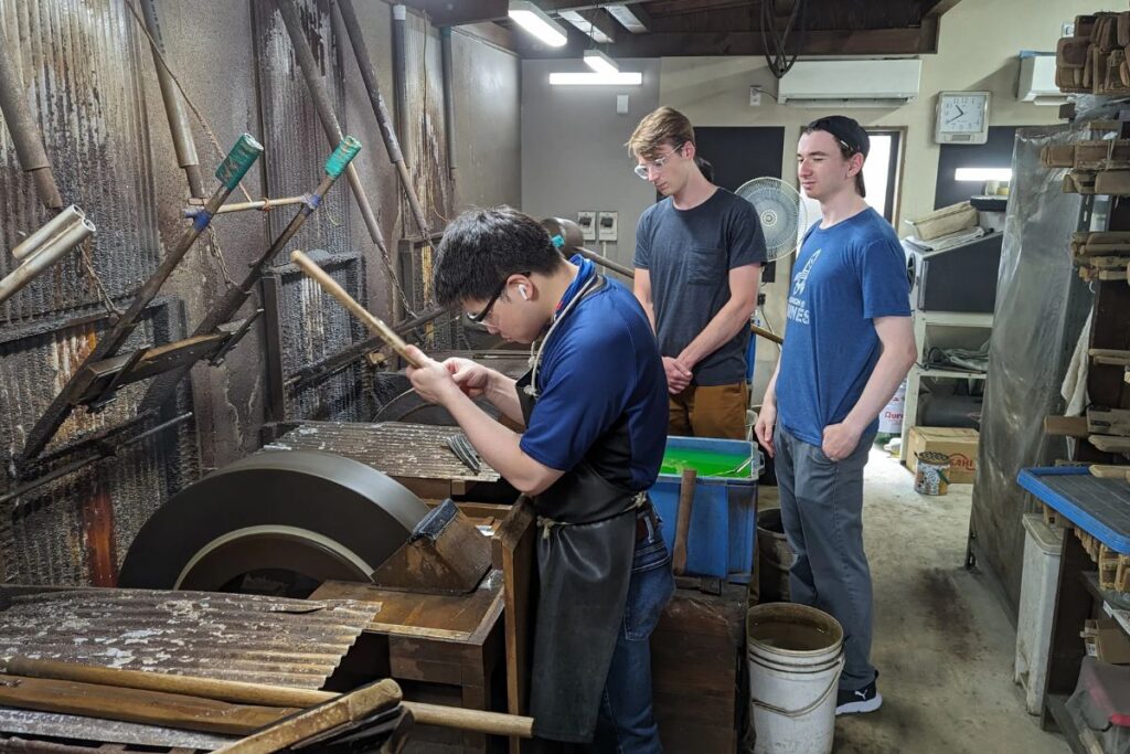 Two visitors watching an artisan craft a steel blade using a large spinning grinding wheel inside a workshop.