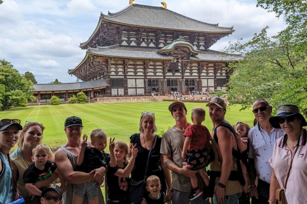 A multi-generational group of travelers, including adults and children in carriers, poses together in front of a giant ancient wooden temple building.