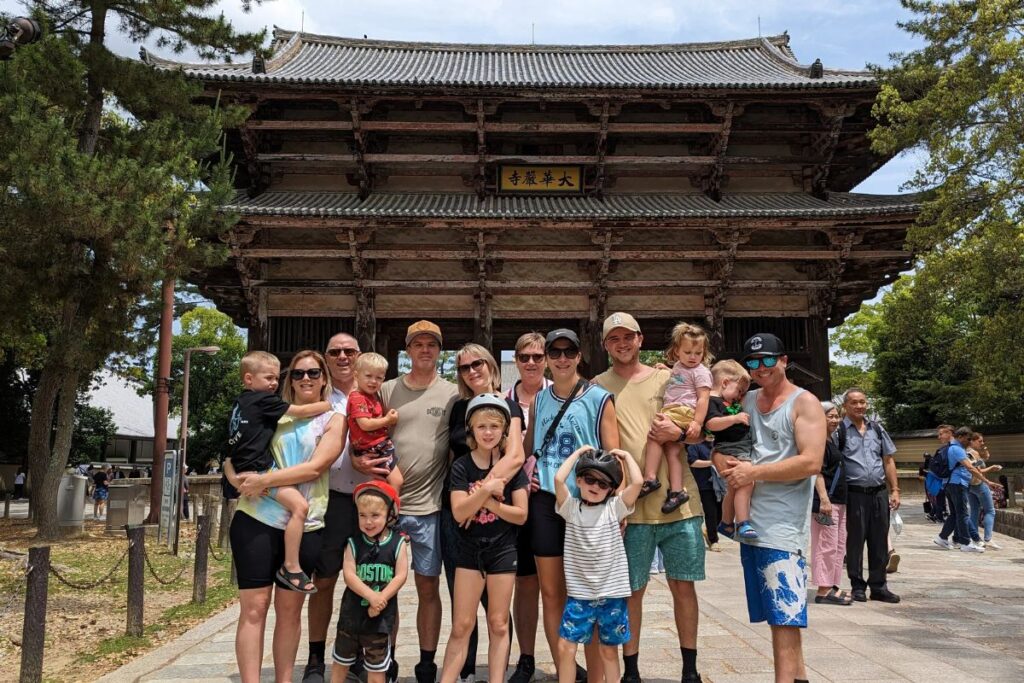 Several adults and children with safety helmets pose in front of a massive historic wooden gate structure.