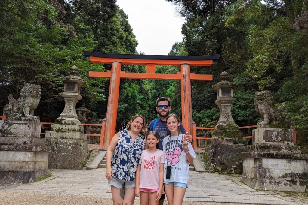 A family of four stands together under a large bright orange shrine entrance gate surrounded by stone lanterns.