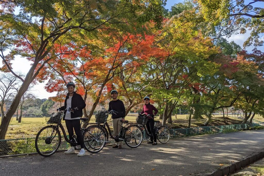Three riders on electric motorized two-wheeled vehicles travel down a paved path lined with colorful autumn maple trees.