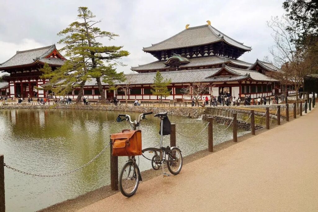A compact folding cycle parked along a scenic pond with a massive traditional Japanese temple in the background on a Brompton bike tour Nara.