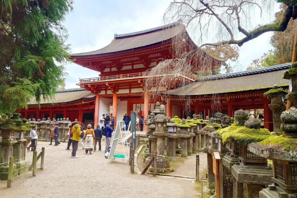 Travelers exploring a bright red ancient shrine entrance surrounded by rows of historic stone lanterns.
