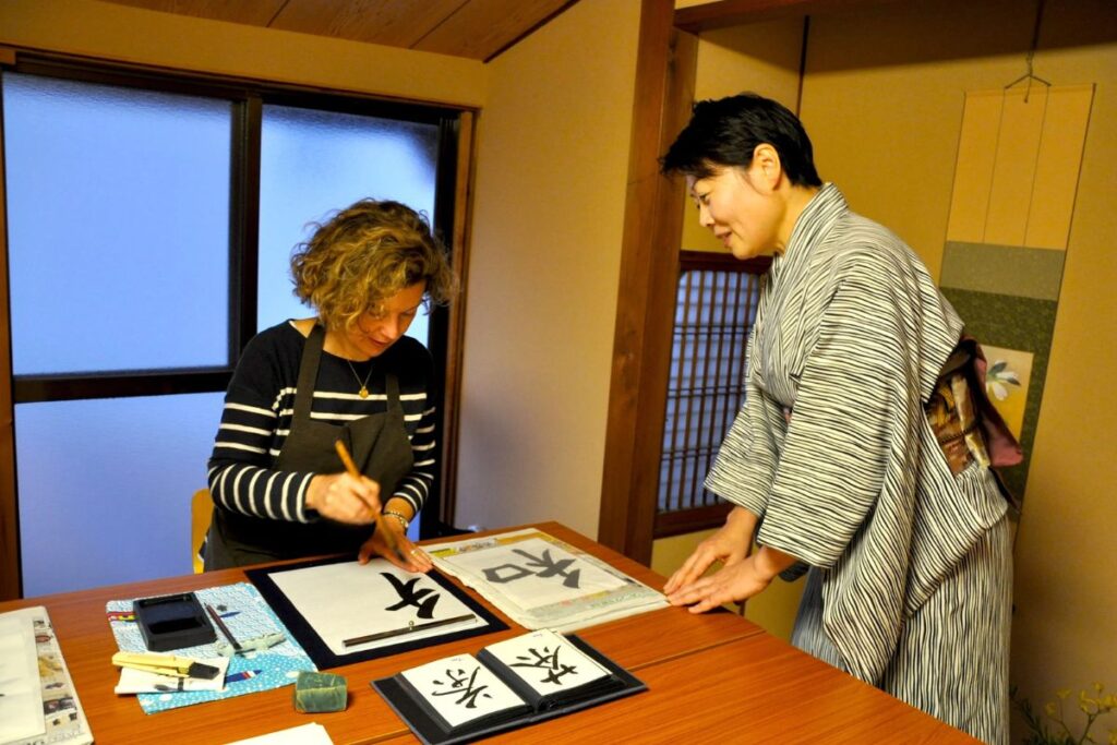 A woman painting Kanji with a brush under the guidance of a kimono-clad instructor on a private calligraphy experience Kyoto.