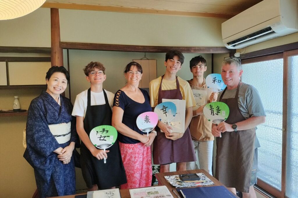 A happy family holding up hand-painted paper fans alongside their Japanese instructor inside a traditional townhouse room.