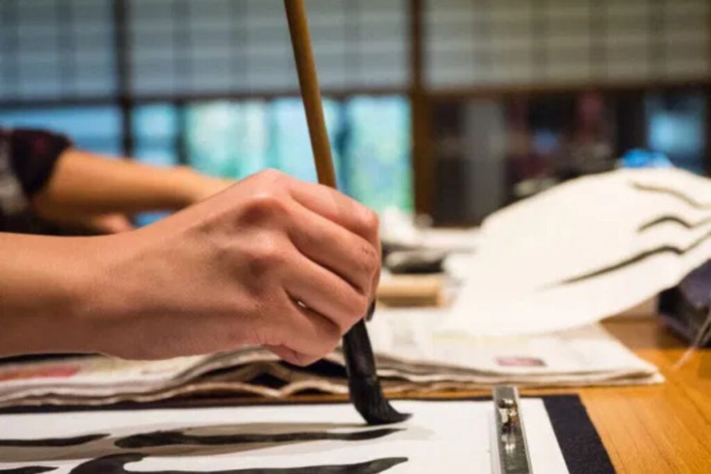 Close-up of a hand carefully guiding a bamboo brush with black ink onto white paper.
