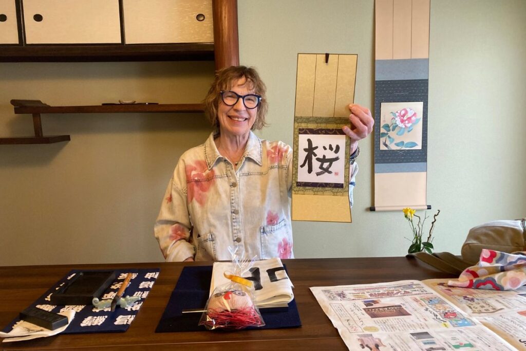An older woman with blue glasses smiling proudly while displaying her finished hanging scroll artwork at a wooden table.