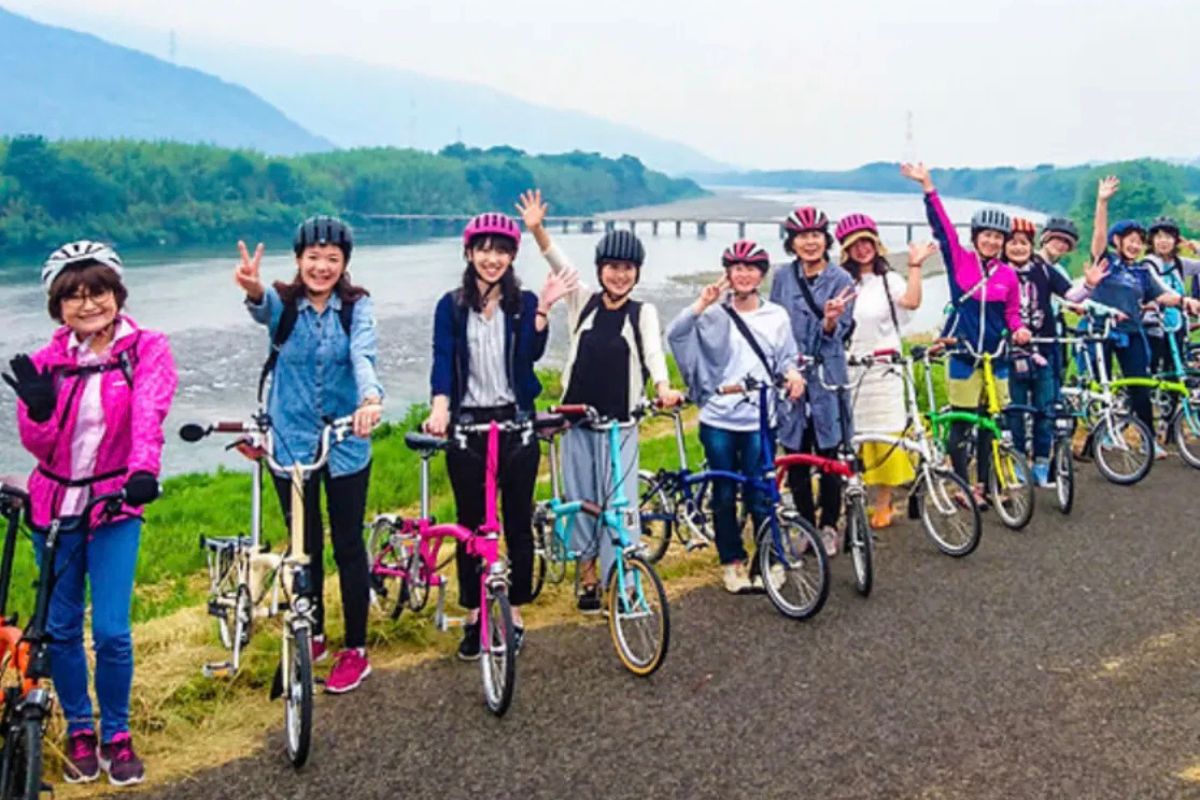 A large group of smiling riders wearing safety helmets waving next to their folding cycles along a scenic river path.