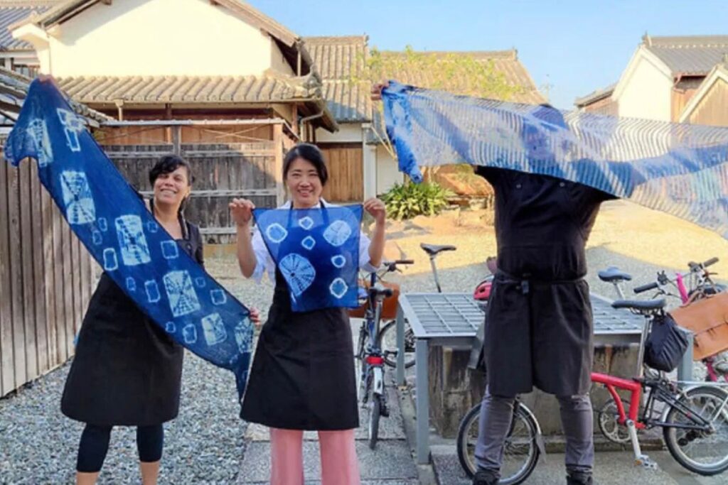 Three happy people holding up freshly dyed blue indigo fabrics outside traditional merchant buildings with folding cycles parked nearby.