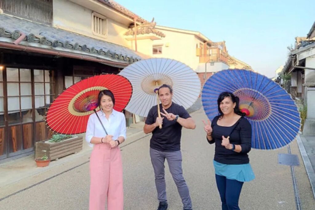 Three travelers smiling and posing with colorful traditional Japanese paper umbrellas in the middle of a historic street lined with old wooden houses.