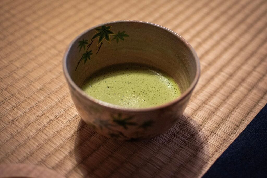 Close-up of a decorative ceramic bowl filled with warm, frothy green matcha resting on a woven tatami mat.