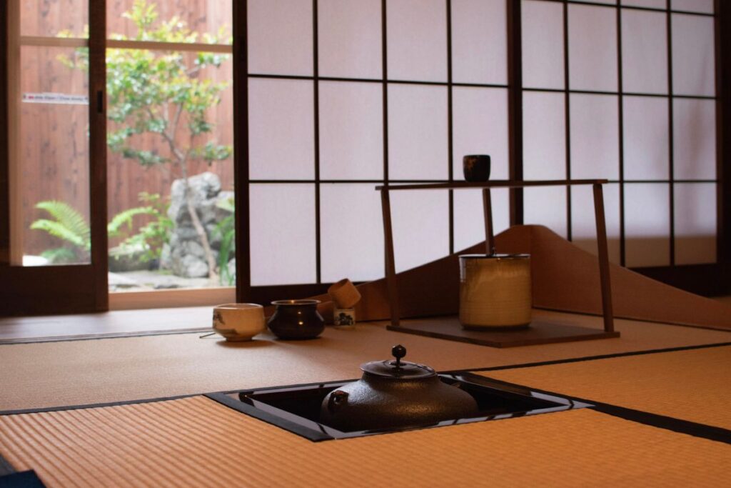 A traditional room featuring woven tatami mats, an iron floor kettle, and sliding doors opening up to a peaceful outdoor garden.
