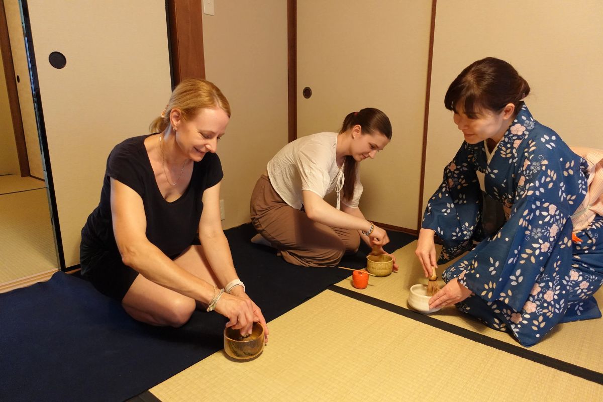 Two women smiling while using bamboo whisks on a tatami mat alongside a local instructor dressed in a kimono.