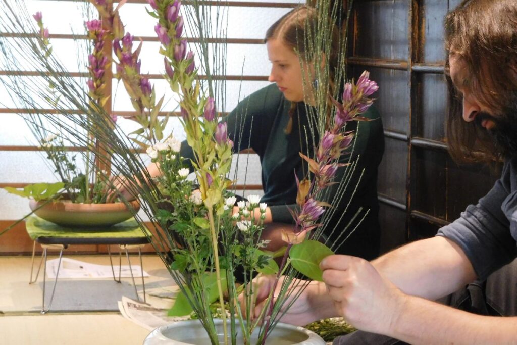 Two focused students carefully placing tall purple blossoms and green stems into shallow ceramic bowls on tatami mats.