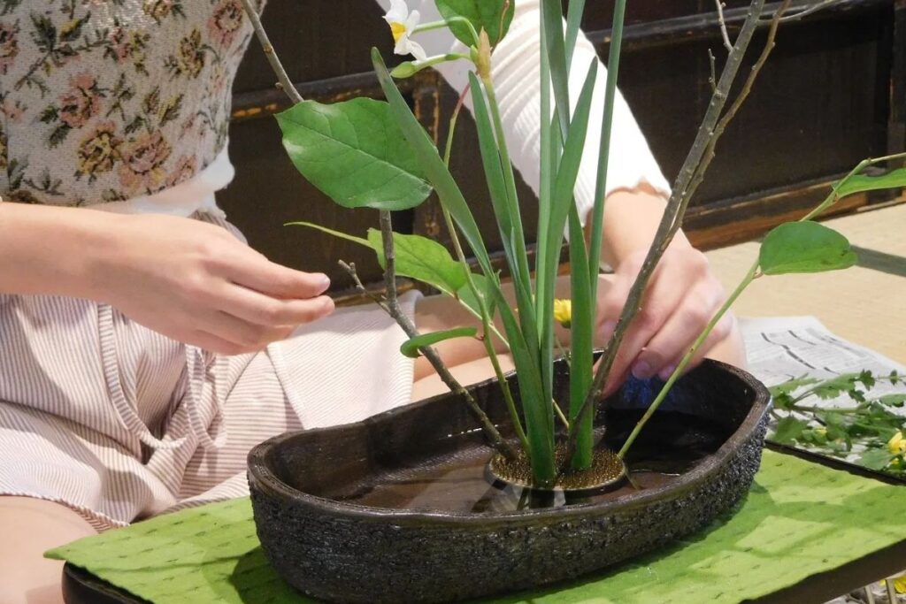 Close-up of a person's hands carefully positioning botanical stems into a pin frog inside a dark textured water basin.