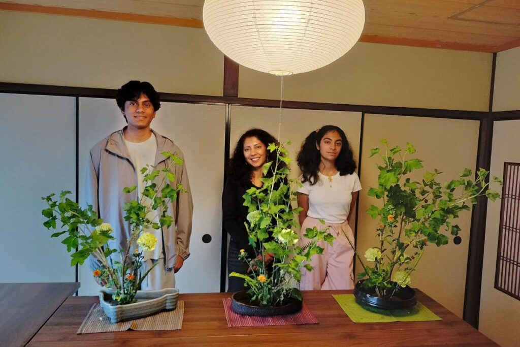 Three people standing proudly behind their finished tall leafy floral designs on a wooden table inside a traditional Japanese room.
