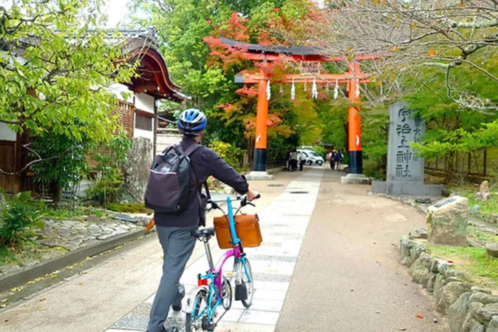A person walking a folding cycle towards a bright orange traditional shrine gate surrounded by colorful autumn foliage.