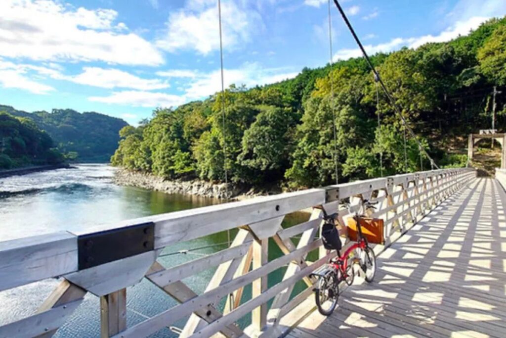 A red folding cycle parked on a white wooden suspension bridge overlooking a wide river and lush green mountains.