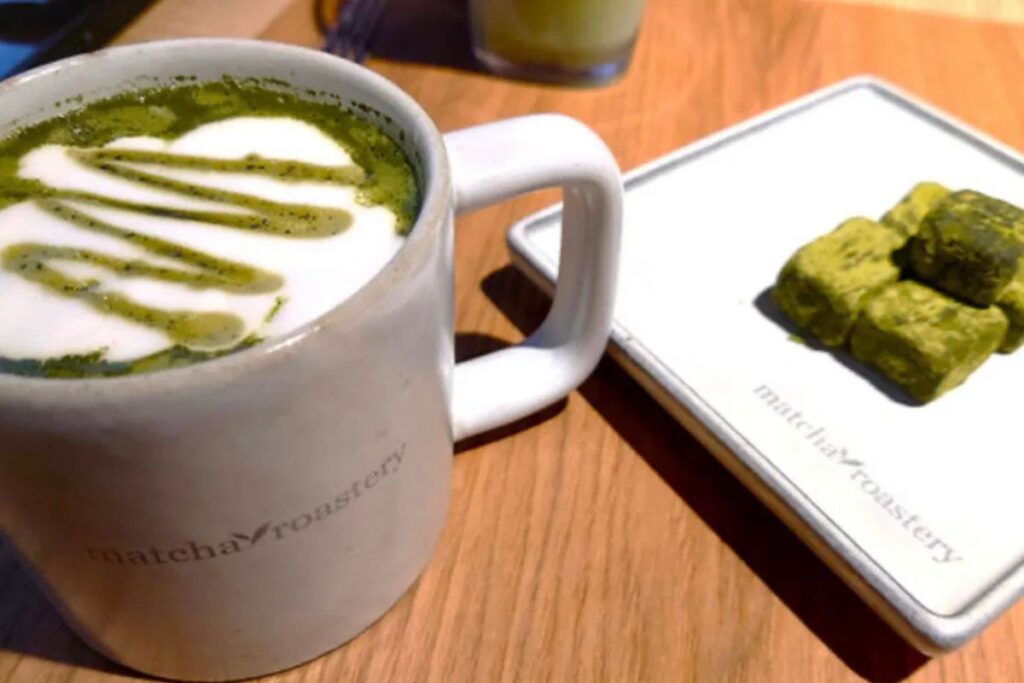 A warm green matcha drink with latte art next to a small plate of square sweet treats on a wooden café table.