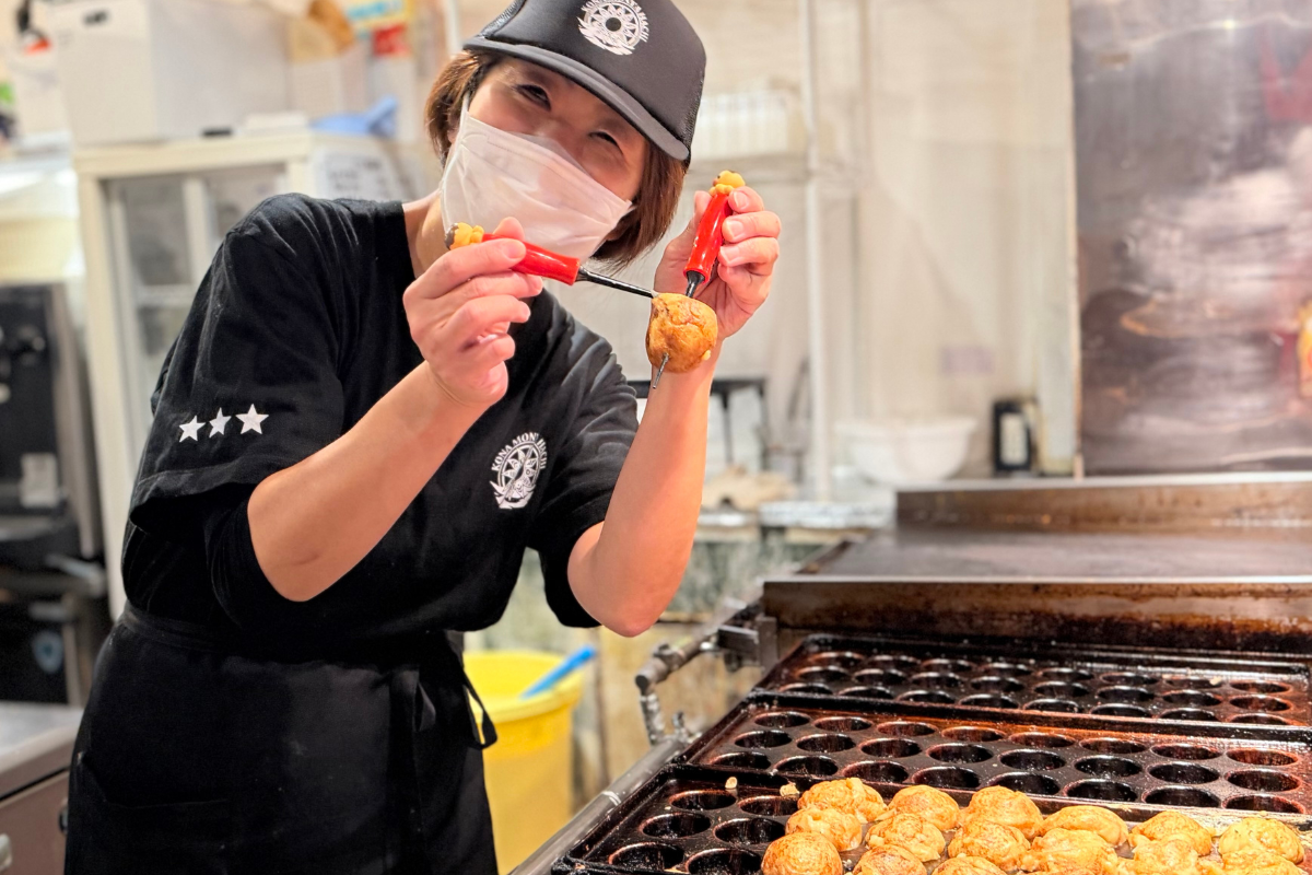 A smiling culinary instructor holding up a freshly cooked, round takoyaki on a skewer over a large commercial griddle plate.