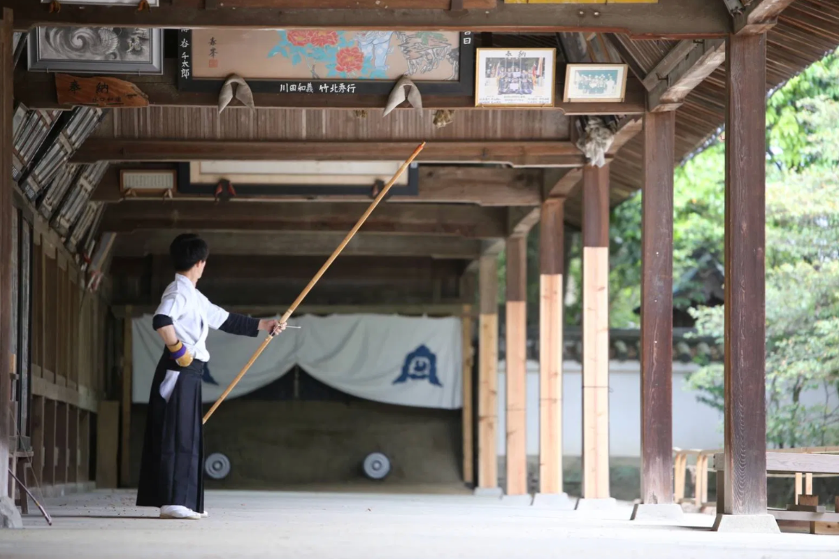 A practitioner dressed in a traditional black and white Hakama stands inside a wooden Shinto shrine corridor holding a large bamboo bow.