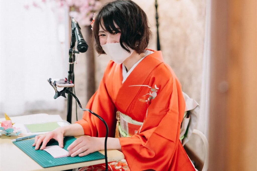 A friendly local host wearing a traditional kimono demonstrating precision paper folding at a desk inside a Tokyo origami private workshop studio.