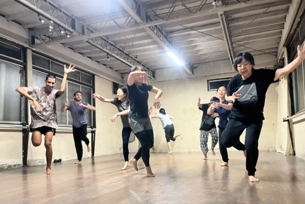 A group of workshop participants barefoot in a dance studio, practicing expressive and mindful Butoh movements with open arms during a guided session.