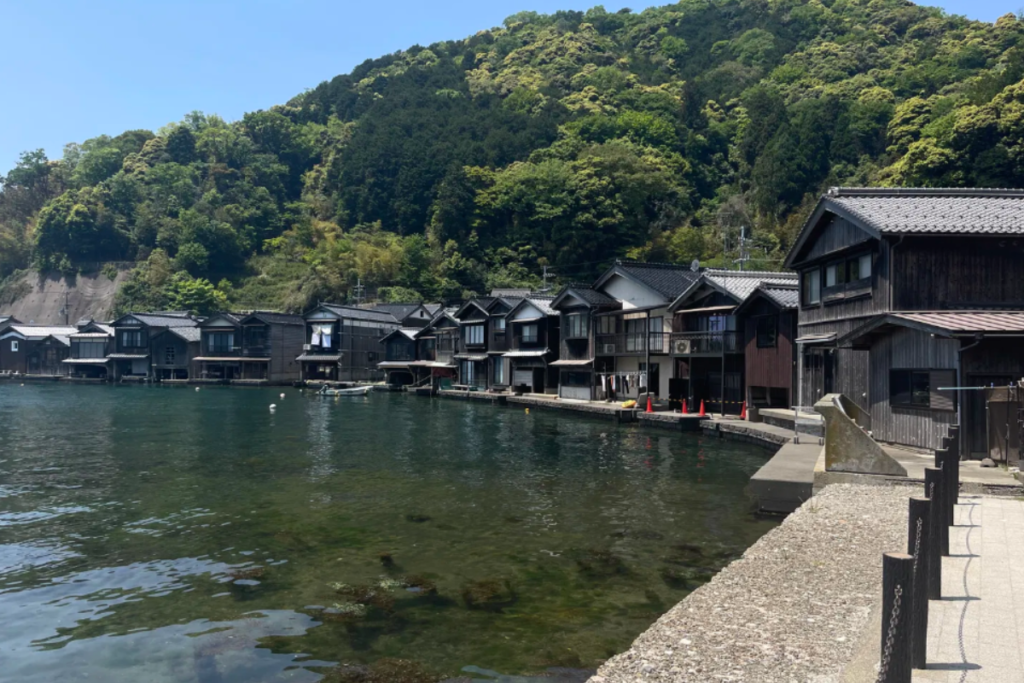 A line of traditional wooden Funaya boathouses built directly over the water’s edge along the green hills of Ine bay.