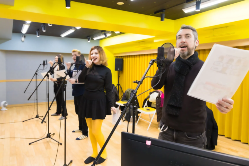 A group of participants stands in front of studio microphones, practicing their lines and expressions during a beginner-friendly voice acting experience.