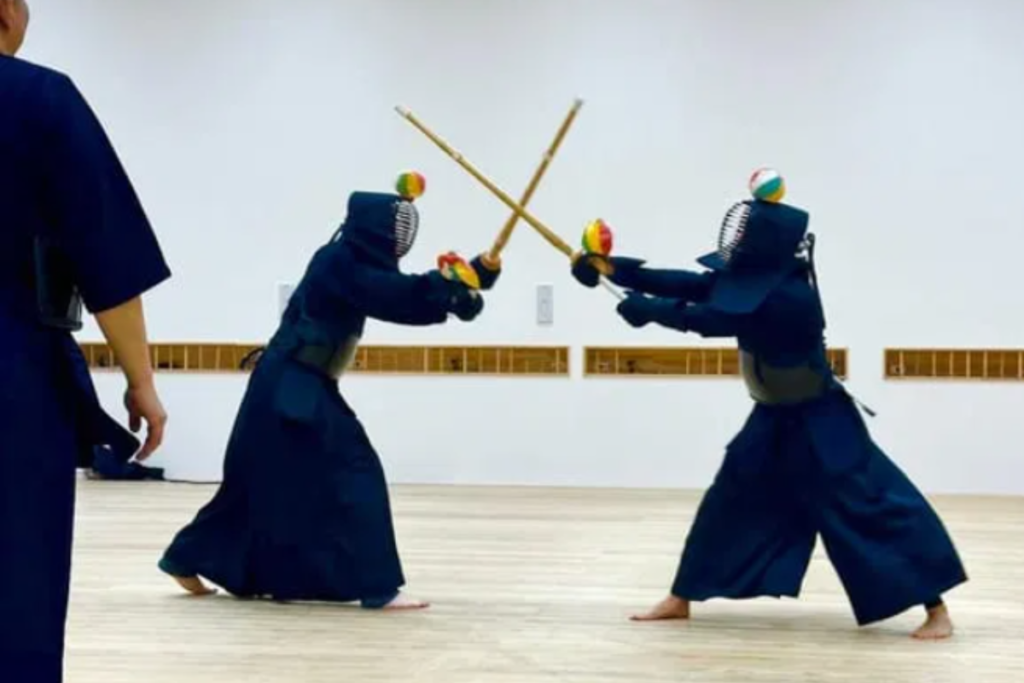Two Kendo practitioners facing each other inside a wooden dojo floor, practicing a striking stance with their bamboo practice swords while wearing full dark protective armor.