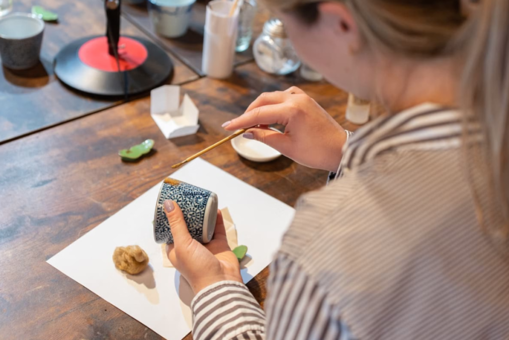 A workshop participant uses a fine brush to carefully apply gold powder accents onto the rim of a blue patterned ceramic cup during a Kintsugi class.