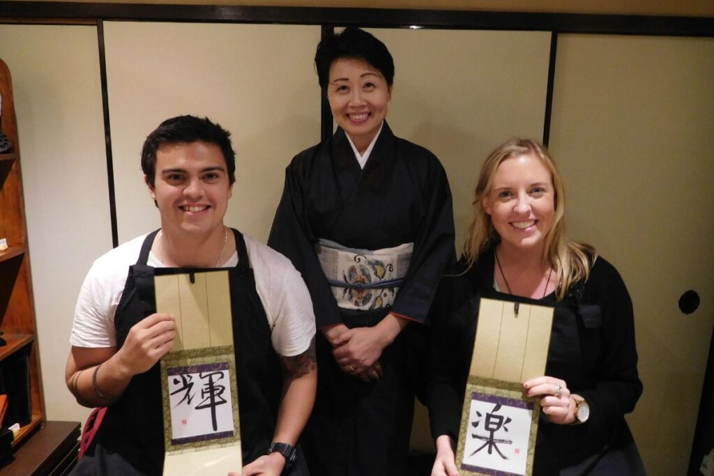 A smiling couple holding up their finished Kanji artwork alongside their instructor during a private calligraphy experience Kyoto.