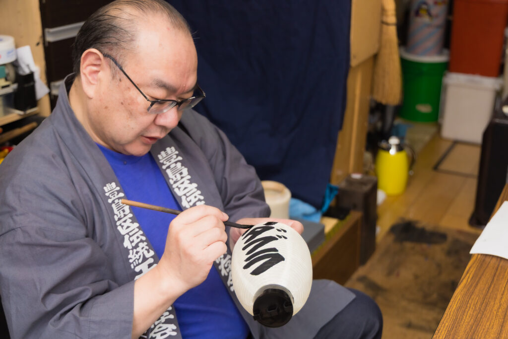 A Tokyo-certified master craftsman with glasses focuses intently on demonstrating painting techniques on a traditional white paper lantern inside his workshop.