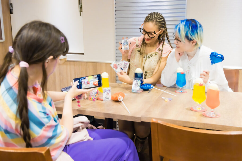 Two smiling women hold up colorful anime character coasters and custom keychains at a table with themed drinks inside an anime collaboration cafe in Tokyo.