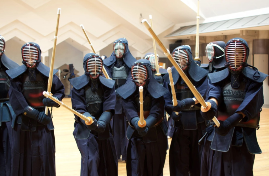 A line of Kendo practitioners standing inside a dojo wearing full protective armor, headgear, and holding bamboo practice swords.