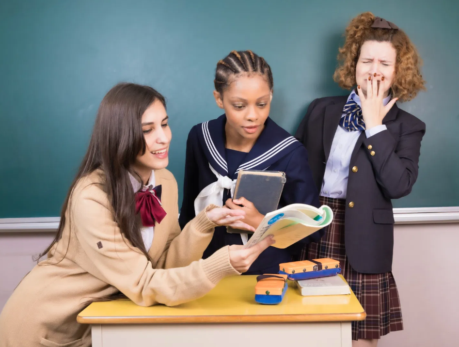 Three young women dressed in traditional Japanese high school uniforms standing together inside a classroom setting, reading a textbook.