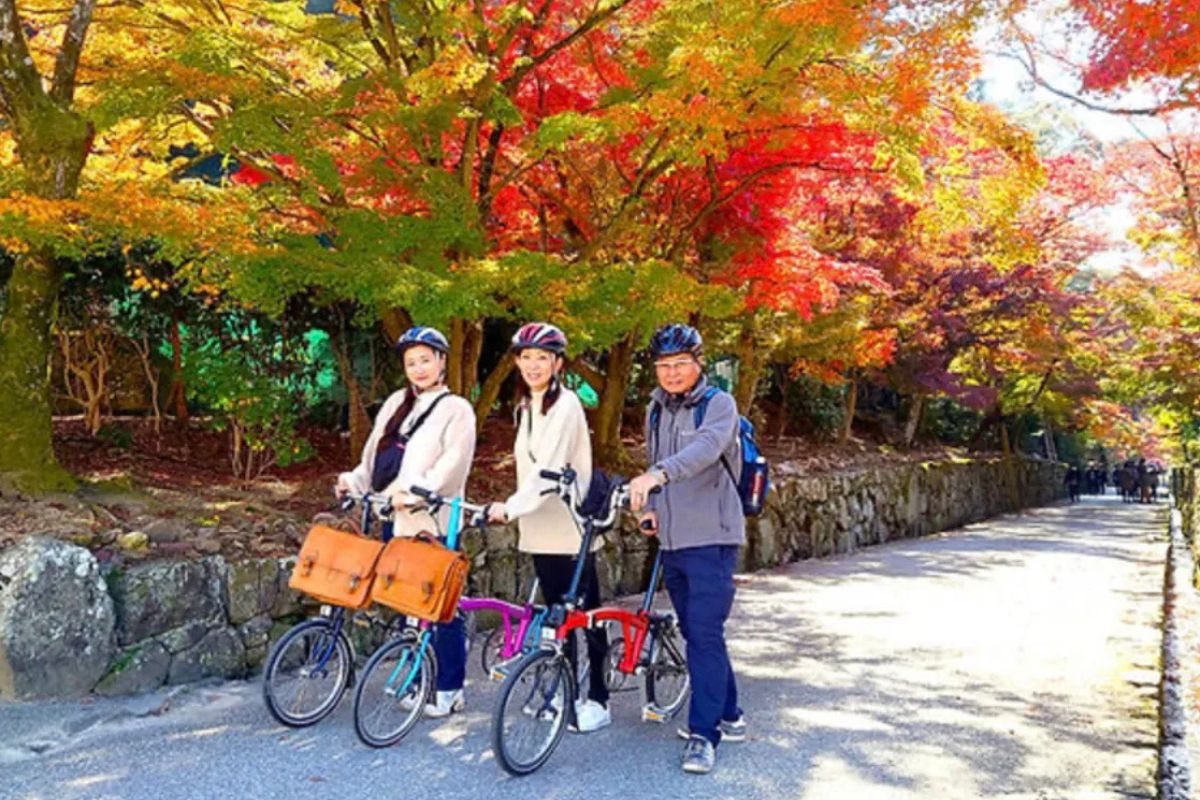 Three smiling travelers posing with their folding cycles under beautiful autumn leaves on an Uji culture bike tour.